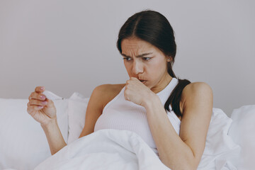 Sick ill tired sad young woman in white tank top sit in bed hold in hand thermometer coughing in bedroom lounge home in own room house wake up. Healthy lifestyle disease treatment cold season concept.