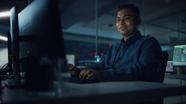 Night Office: Portrait of Handsome Indian Man in Working on Desktop Computer. Digital Entrepreneur Typing, Creating Modern Software, e-Commerce App Design, Programming. Successful Smiling Man