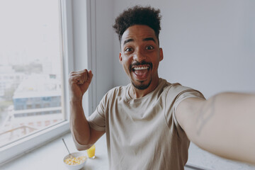 Close up young african american man in casual clothes do winner gesture clench fist do selfie shot pov on mobile phone prepare cooking food in light kitchen at home alone indoor Healthy diet concept