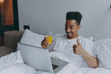 Young african american man in nightwear hold work use laptop pc computer talk video call show thumb up drink juice lying in bed rest relax spend time in bedroom lounge home in own room house wake up.