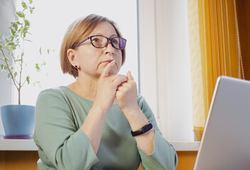 Thoughtful mature woman in eyeglasses with laptop, looking in distance, thinking over task, analyzing solutions, options solving problem, feeling doubtful, sitting indoor. Making decision