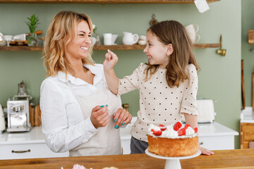 Happy chef cook baker mom woman in white shirt work with smiling child baby girl helper play with mum at kitchen table home. Cooking food process concept Mommy little kid daughter prepare fruit cake
