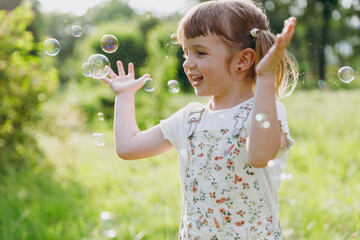 Little smiling excited happy kid girl 5-6 years old in white casual clothes blowing bubbles play on park green sunshine lawn, spending time outdoor in village countyside during summer time vacations.