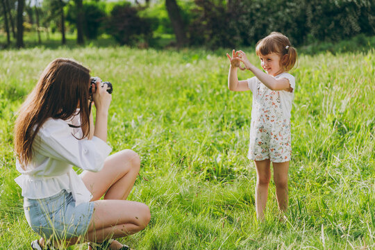 Photographer young woman wear white clothes take picture shot on photo camera of child baby girl 5-6 years old show heart-shape sign Mommy rest with little kid daughter outdoor. Love family concept