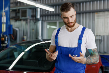 Pensive young professional technician car mechanic repairman man wear denim blue overalls white...