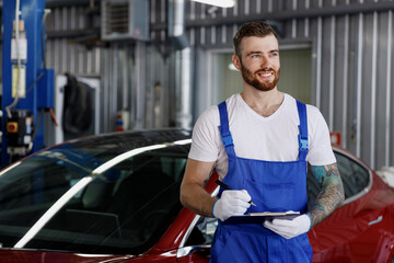 Young minded male professional car mechanic man 20s wears denim blue overalls white t-shirt gloves write hold clipboard with papers document work in light modern vehicle repair shop workshop indoors