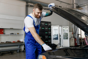 Strong smiling happy confident young male professional technician car mechanic man wears denim blue overalls white t-shirt raised hood bonnet work in light modern vehicle repair shop workshop indoors.