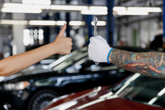 Close up cropped photo of professional technician car mechanic male hands in gloves hold wrench key tool, female driver owner arm show thumb up gesture in modern vehicle repair shop workshop indoor. - Powered by Adobe