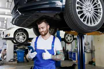 Young professional technician mechanic man in blue overalls t-shirt use hold mobile cell phone show thumb up stand near car lift check technical condition work in vehicle repair shop workshop indoors.