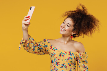 Young happy smiling beautiful woman 20s with culry hair wearing casual clothes doing selfie shot on mobile cell phone post photo on social network isolated on plain yellow background studio portrait