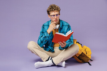 Full body young boy teen student wear casual clothes backpack headphones glasses sit reading book isolated on violet background studio portrait. Education in high school university college concept