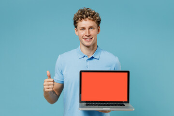 Young curly man 20s years old wear azure t-shirt show thumb up like gesture hold use laptop pc computer with blank screen workspace area isolated on plain pastel light blue background studio portrait