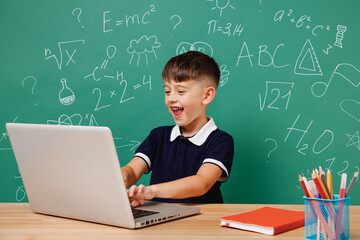 Young happy satisfied male kid school boy 5-6 years old in t-shirt sit use laptop pc cocmputer isolated on green wall chalk blackboard background Childhood children kids education lifestyle concept