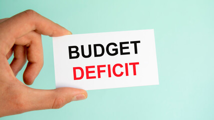 businessman's hand holding paper business card with text budget deficit, closeup light blue background