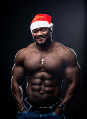 Fototapeta na wymiar Happy New Year. Full length view of the sexy muscular man wearing Christmas hat standing pensively with naked torso. Naked bodybuilder posing for camera at studio
