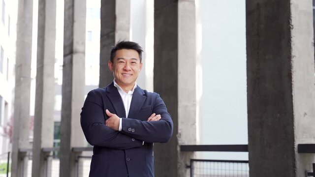 Portrait happy friendly asian businessman standing on urban city street background outside. Business man office worker arms crossed in formal suit near modern office building looking at camera smiling