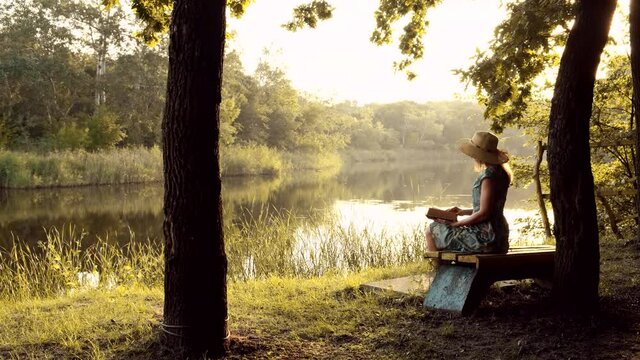 Beautiful young woman in dress and hat sitting on bench under tree and reading a book at sunset. Lady with book resting relaxing on nature near lake or river outdoors. Enjoying vacation. Girl sitting 