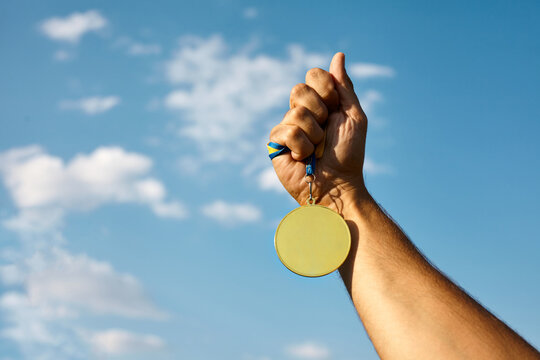 Winner hand raised and holding gold medal with ribbon against blue sky.