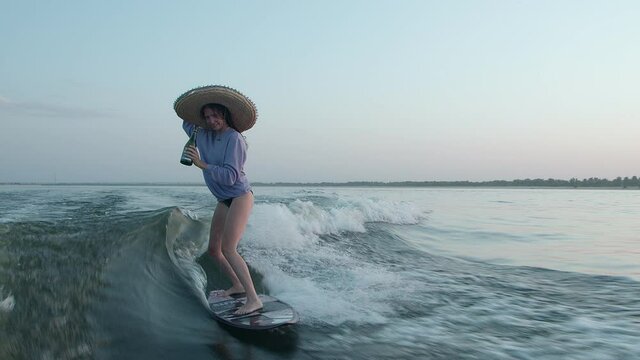 A surfer in a straw hat jumps on a wakeboard with champagne and a phone in her hands. An experienced wakeboarder sprays water drops into the camera.