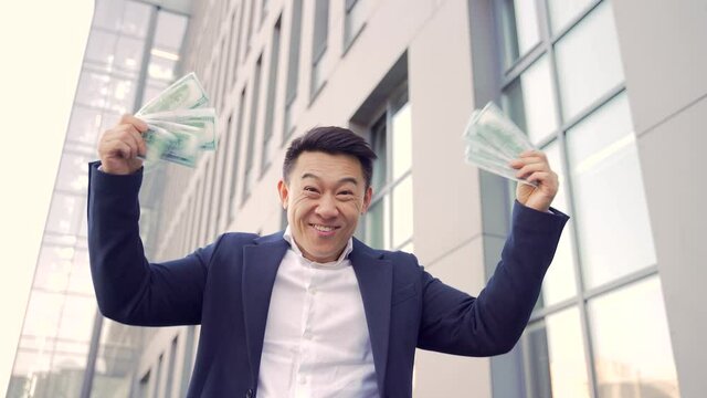 Happy successful asian businessman counts, waves, throws, money background a modern office building looking at camera in downtown. Winner success in betting stock market celebrating victory. outside