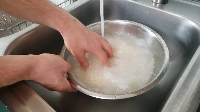 Home cooking - Washing while white rice in stainless steel bow under running tap water in the sink.