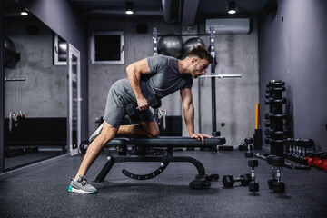 Man doing exercise with dumbbell leaning on sports bench in the gym. Photo of a sexy muscular man in sportswear and good physique on grey background. Strength and motivation, sport, fitness goal - Powered by Adobe