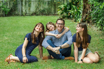Fototapeta premium A family of a mother and father and two daughters sitting outside in the grass of their backyard witht the youngest daughter hugging dad from behind