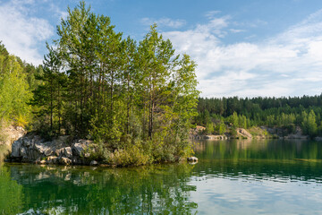 Rocky island in the middle of a blue-green lake with reflective sky and clouds, forest on the shore. Beautiful natural landscape.