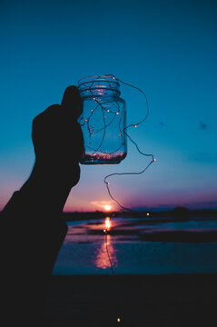 Silhouette of person holding a glass jar with string light in it during sunset on seashore