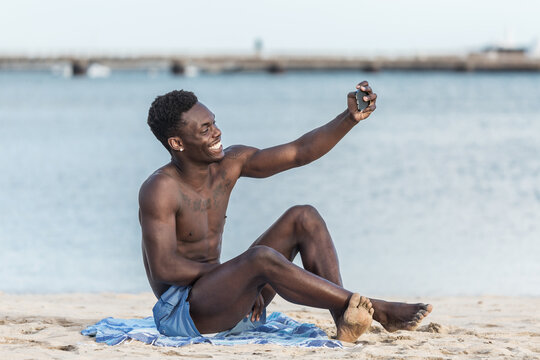 Topless man sitting on beach taking selfie with his cellphone