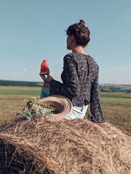 Woman holding a slice of watermelon sitting beside a straw hat with flowers in it on a hay in grass field
