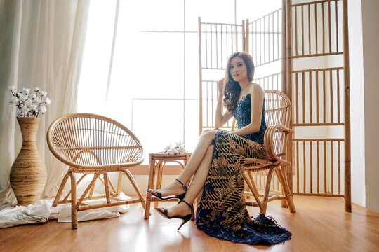 Woman in a long designer dress and high heels sitting on a wooden chair beside a window indoors during daytime