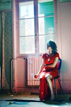 Woman in red dress sitting on chair next to window smoking