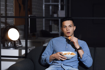 Handsome young man eating tasty nachos while watching TV at home late in evening