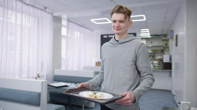 Portrait of positive young man posing with dessert on tray in canteen indoors. Happy smiling Caucasian male student looking at camera standing in cafeteria with sweet lunch