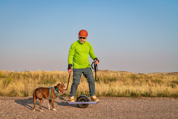 mature senior male is riding one-wheeled electric skateboard with a pitbull dog on a prairie road,...