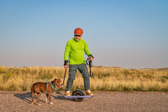 Mature Senior Male Is Riding One-wheeled Electric Skateboard With A Pitbull Dog On A Prairie Road, Late Summer Scenery With Haze And Smoke From Distant Wildfires