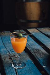 orange carrot drink cocktail in gold rimmed wine glass with green olive garnish on blue wooden pallet table 