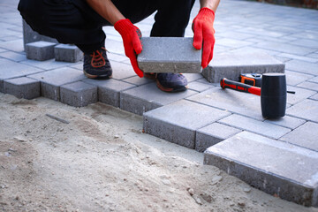 The master in yellow gloves lays paving stones in layers. Garden brick pathway paving by professional paver worker. Laying gray concrete paving slabs in house courtyard on sand foundation base.