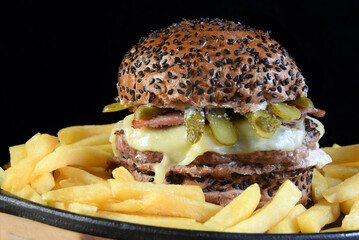 beef burger sandwich with bacon, pickles, cheese, french fries and dark bun isolated on black background.