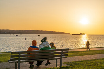 Family sitting on the bench, enjoy the sunset time at English Bay Beach, Vancouver City beautiful landscape. Concept of happiness and peaceful.