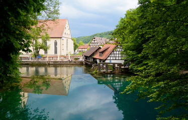a scenic view of emerald-green lake Blautopf and the historic old hammer mill, Blaubeuren Abbey, and timber-framed houses in the background in the old romantic German town of Blaubeuren (Germany)