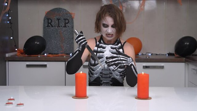 Frightening witch with creepy makeup in skeleton costume heats knife in candlelight and pierces balloon to perform sinister ritual. Show at Halloween party.