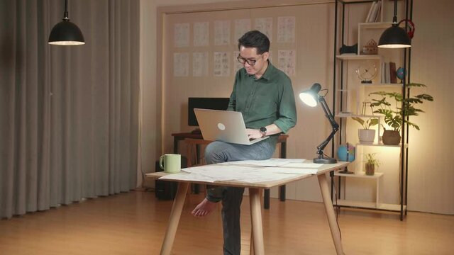 Creative Designer Sits On His Desk Holds Laptop On The Knees And Working On The Project . Creative Designe And Gaming Content Development Studio
