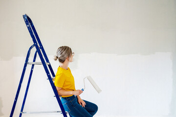 A woman in a yellow T-shirt is painting a wall in her house. Girl sitting on a stepladder near a...