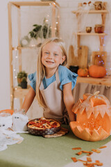 a blonde baby girl with long hair in an apron in the kitchen, decorated with pumpkins and garlands for Halloween, prepares a focaccia pie. space for text. High quality photo