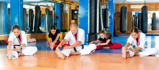 Group of young sporty people wearing kimonos stretching during warming up before martial arts training in gym .