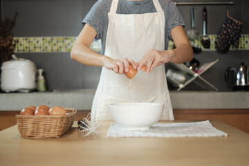 Close-up front view footage, a female cook in a white apron is cracking an egg into a cup to prepare a meal on a wooden table in the home's kitchen. Eating egg yolks is a healthy breakfast.