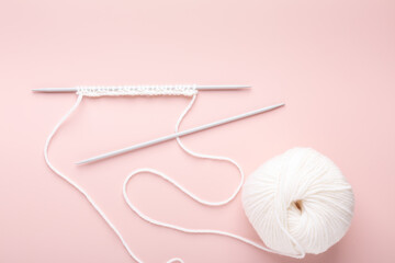Knitting project in progress. White knitting wool and knitting needles on pastel pink background. Female hobby knitting. Top view