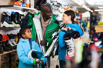 Fototapeta na wymiar Positive young African man and European woman with preteen son choosing together ski boots for skiing in store of sports gear